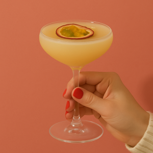 Main tenant un verre à cocktail Pornstar Martini garni d’une demi-passion, sur fond corail.