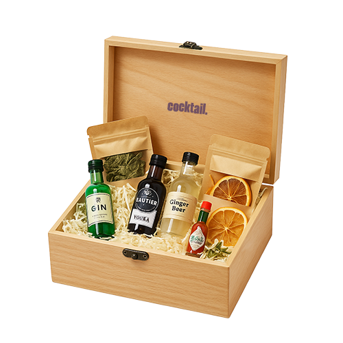 Boîte en bois ouverte sur fond blanc, contenant plusieurs mini bouteilles d’alcool, flacons de jus, sachets d’épices et de fruits séchés, présentés sur du papier frisotté blanc.