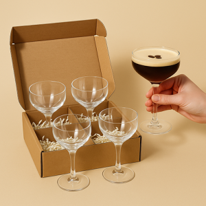 Boîte en carton ouverte contenant trois verres à cocktail debout dans des compartiments séparés avec papier blanc, une case vide et à côté une main tenant un verre rempli d’Espresso Martini.