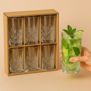 Boîte en carton kraft ouverte contenant 4 verres long drink taillés style cristal avec papier frisotté, et une main tenant un verre rempli de Mojito avec glace, menthe et rondelles de citron vert.