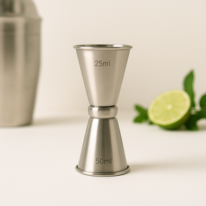 Jigger en inox brossé avec gravure des mesures 25 ml et 50 ml, posé sur fond blanc, accompagné d’un shaker en arrière-plan flouté et d’un demi-citron vert avec feuilles de menthe.