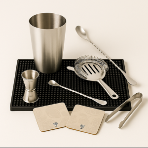 Kit de bar en inox avec shaker Boston, jigger, passoire Hawthorne, cuillère de bar, pince à glaçons, présentés sur un tapis silicone noir, fond blanc.