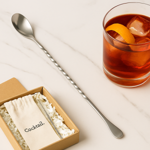 Cuillère de bar inox longue et torsadée posée sur plan de travail marbré, à côté d’un cocktail Negroni et d’une boîte cadeau contenant un pochon marqué « Cocktail. ».