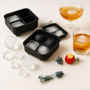 Quatre moules à glaçons en silicone noir présentés sur fond blanc, accompagnés de glaçons démoulés en sphères et cubes, entourés de tranches d’agrumes séchés.